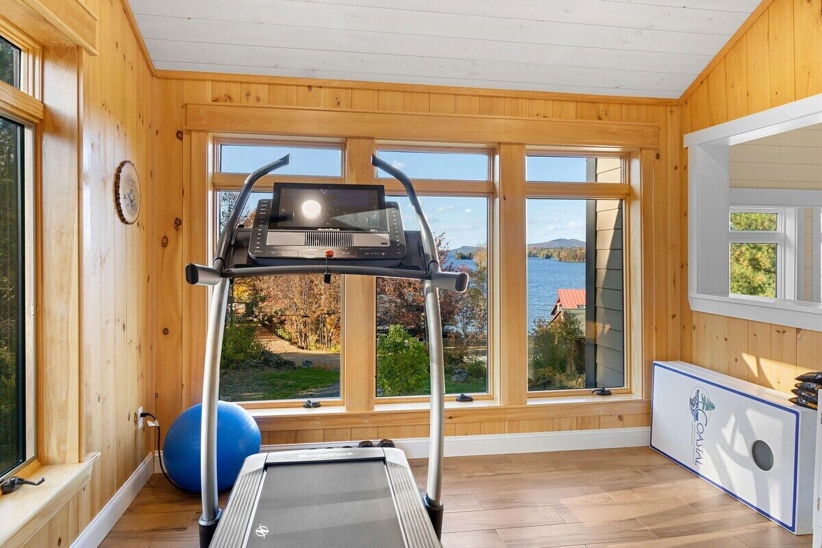 A room with a treadmill, exercise ball, and large windows with a view of a lake.
