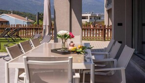 Un espace extérieur aménagé pour les repas, avec une table et des chaises en verre, un vase rempli de fleurs et une vue sur les montagnes en arrière-plan.