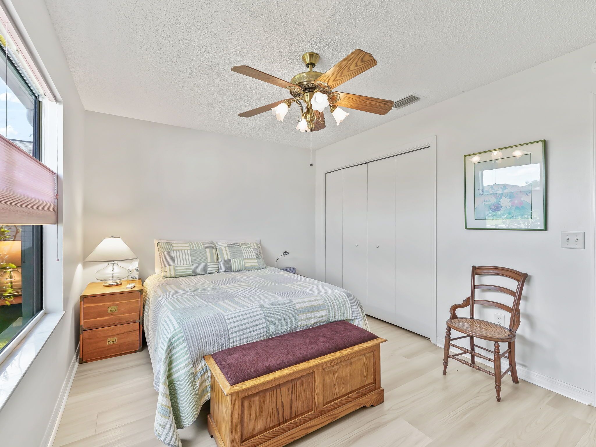 A bedroom with a bed, a wooden bench, a chair, a nightstand with a lamp, a ceiling fan, a wardrobe, and a window with blinds.
