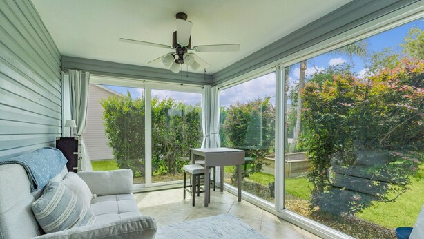 A sunroom with a ceiling fan, a gray sofa with striped pillows, a small table, and a view of a garden.