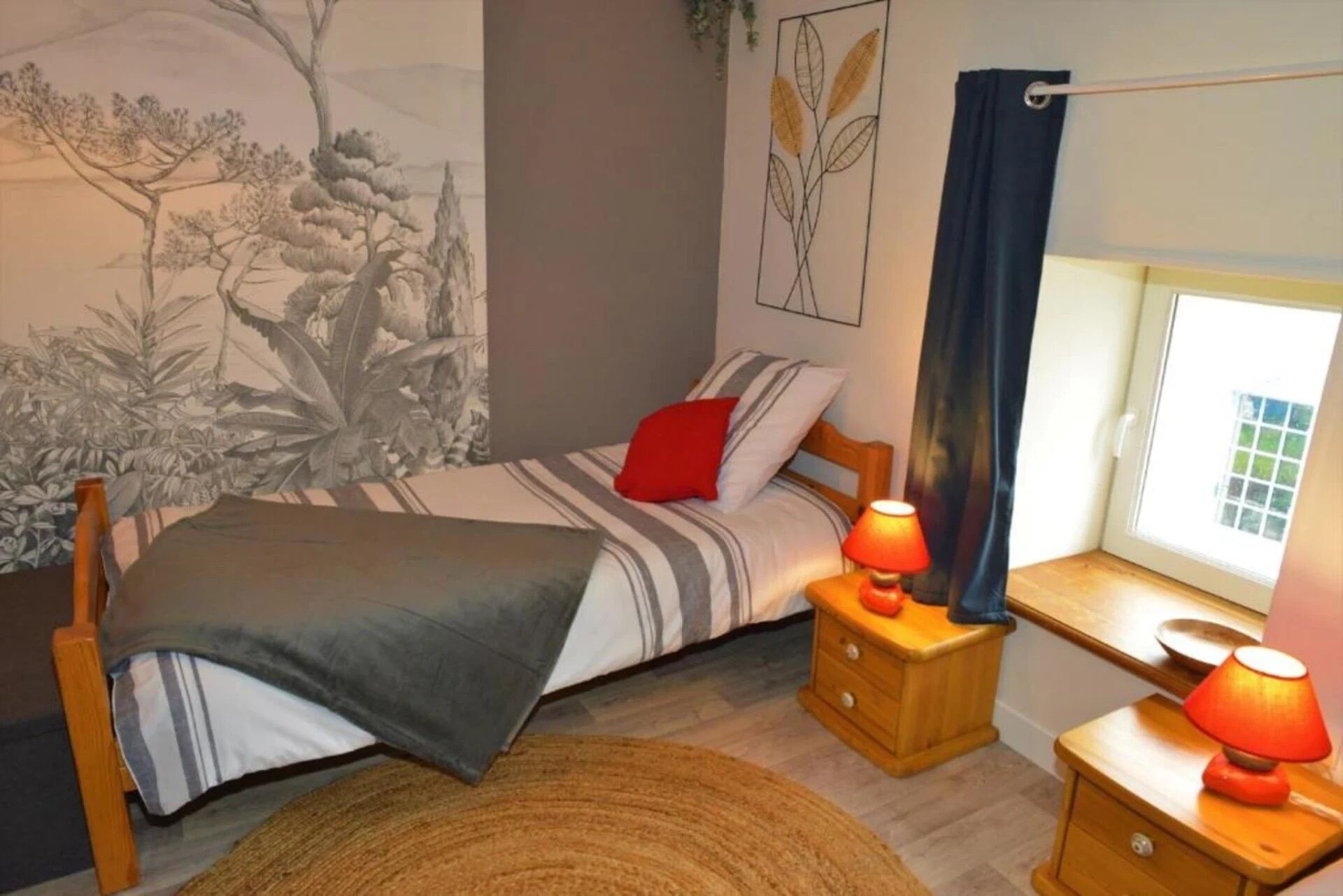 Een slaapkamer met een bed, nachtkastje met lamp, een houten dressoir, een raam met gordijnen, en een muurschildering.