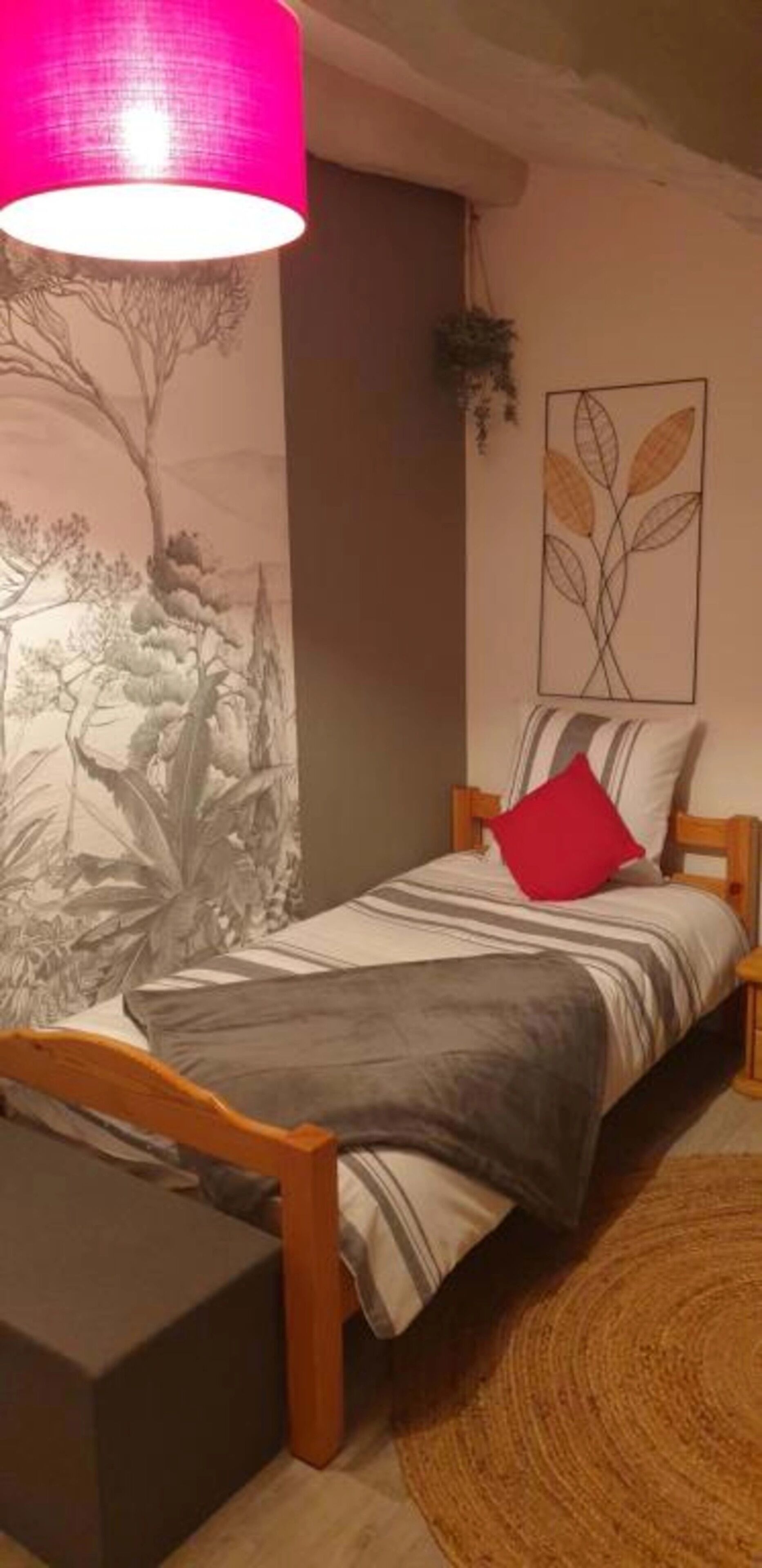 A small, cozy bedroom with a bed, a nightstand, a lamp, and a wall with a nature-themed mural.