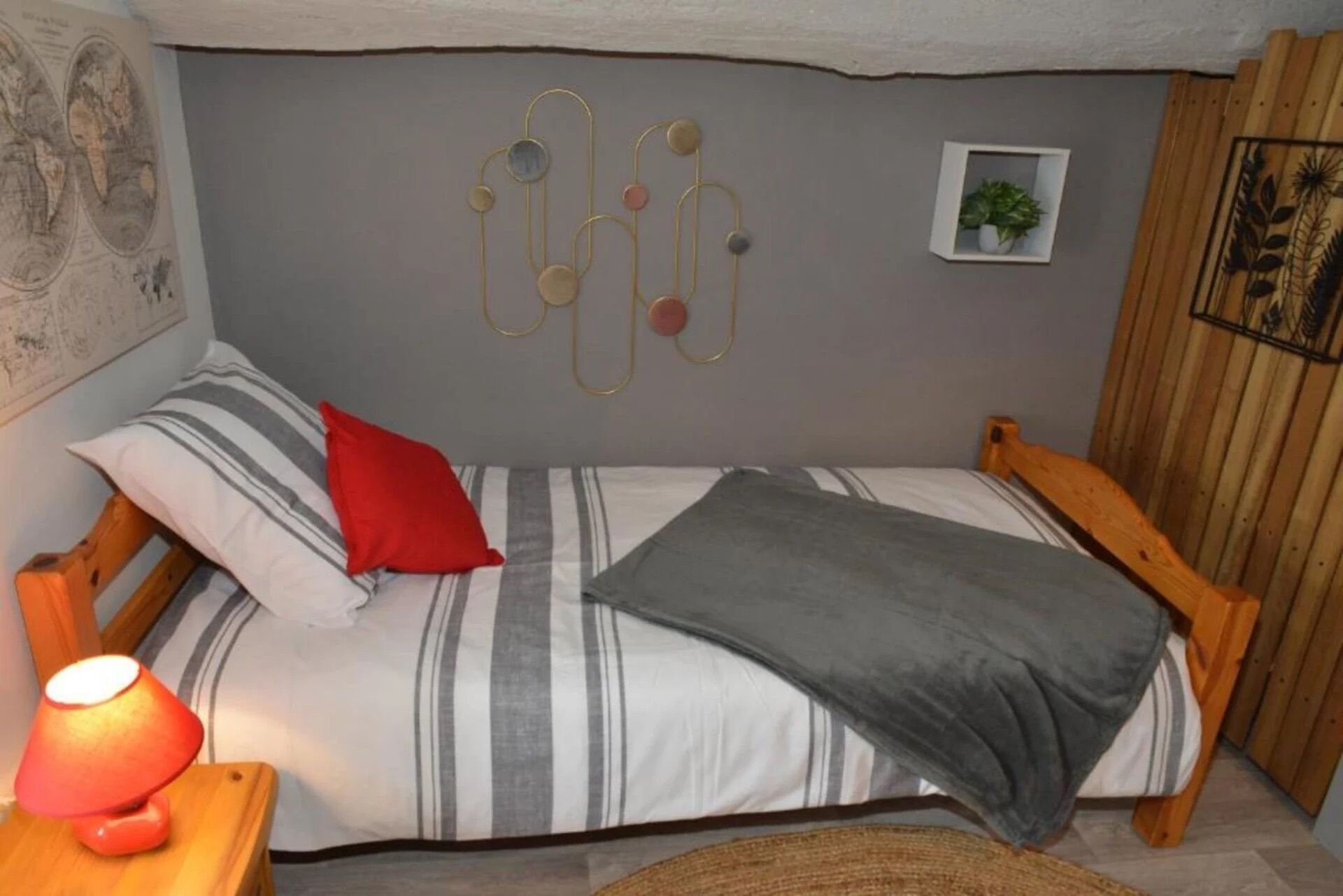 Un dormitorio pequeño y acogedor con una cama individual, ropa de cama a rayas, una almohada roja, una manta gris, una lámpara de pared y una planta pequeña.