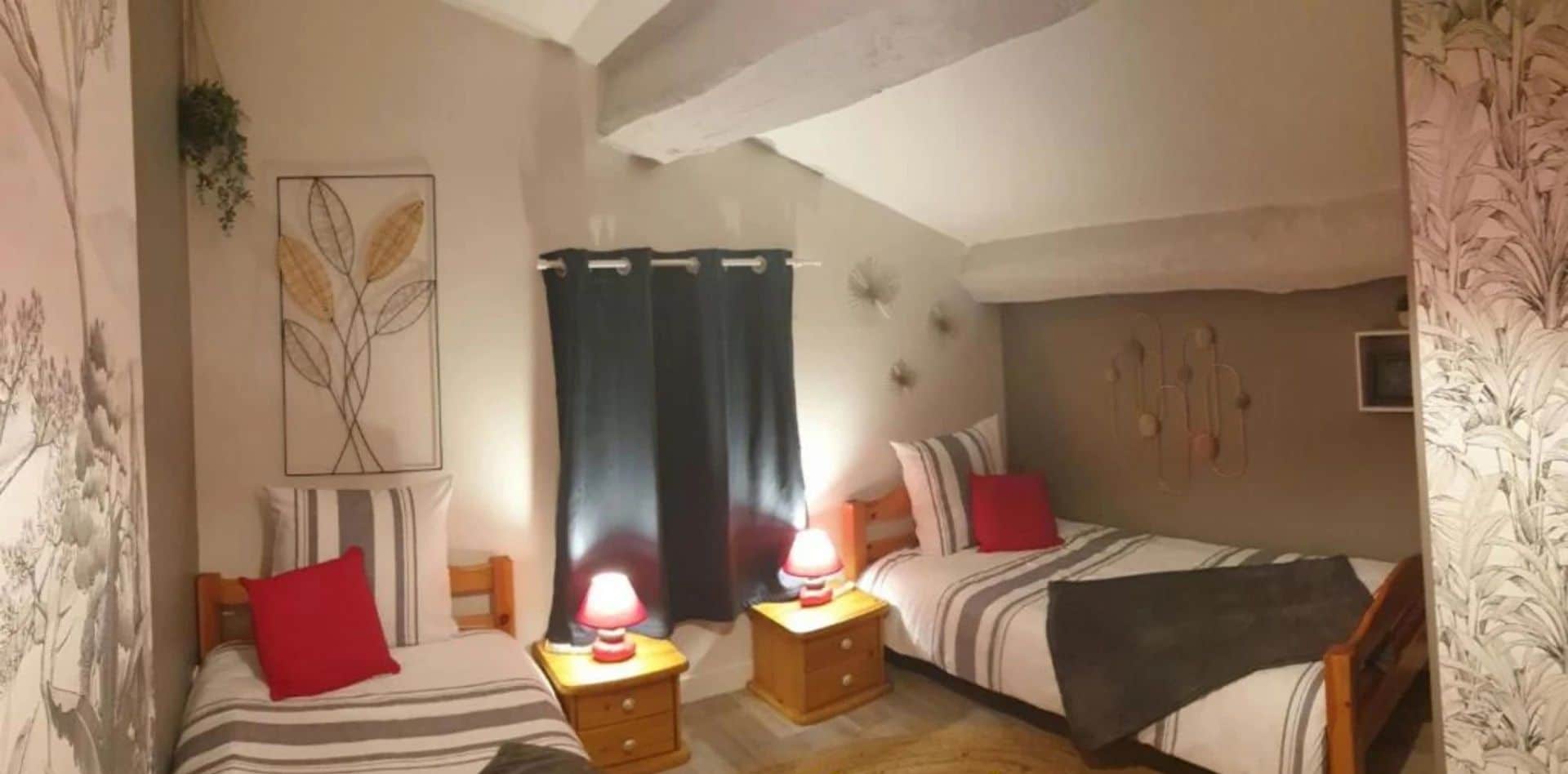 A double room with two beds, each with striped bedding and red pillows. There are bedside tables with lamps, a wall-mounted TV, and a ceiling with exposed beams.