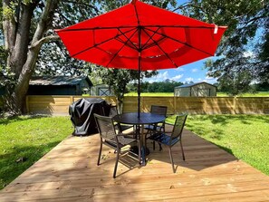 Une terrasse en bois avec une table ronde et des chaises, un parasol rouge et un barbecue.
