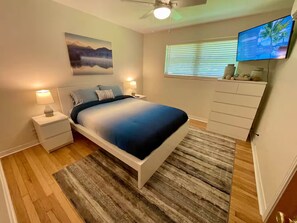 Une chambre à coucher comprenant un lit, des tables de chevet, un téléviseur, un ventilateur de plafond, un tableau et une fenêtre avec des stores.