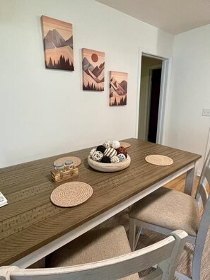 Une table à manger avec un plateau en bois, entourée de quatre chaises à l’assise de couleur claire. Sur la table, on trouve un bol rempli d’objets décoratifs et deux sets de table tissés. Trois tableaux représentant des paysages de montagne sont accrochés au mur au-dessus de la table.