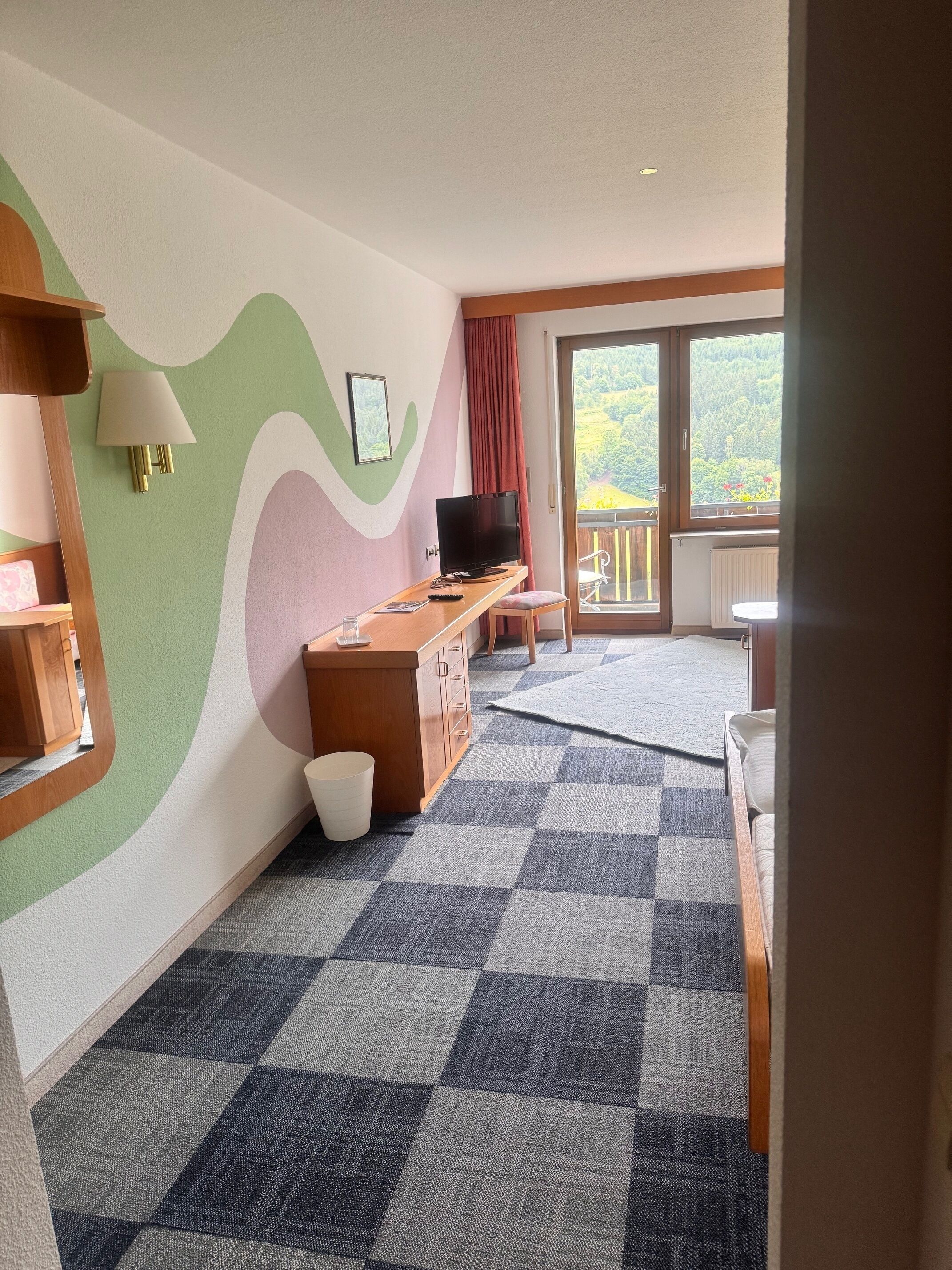 A room with a checkered carpet, a wooden desk with a television, a wall-mounted lamp, and a window with a view of greenery.