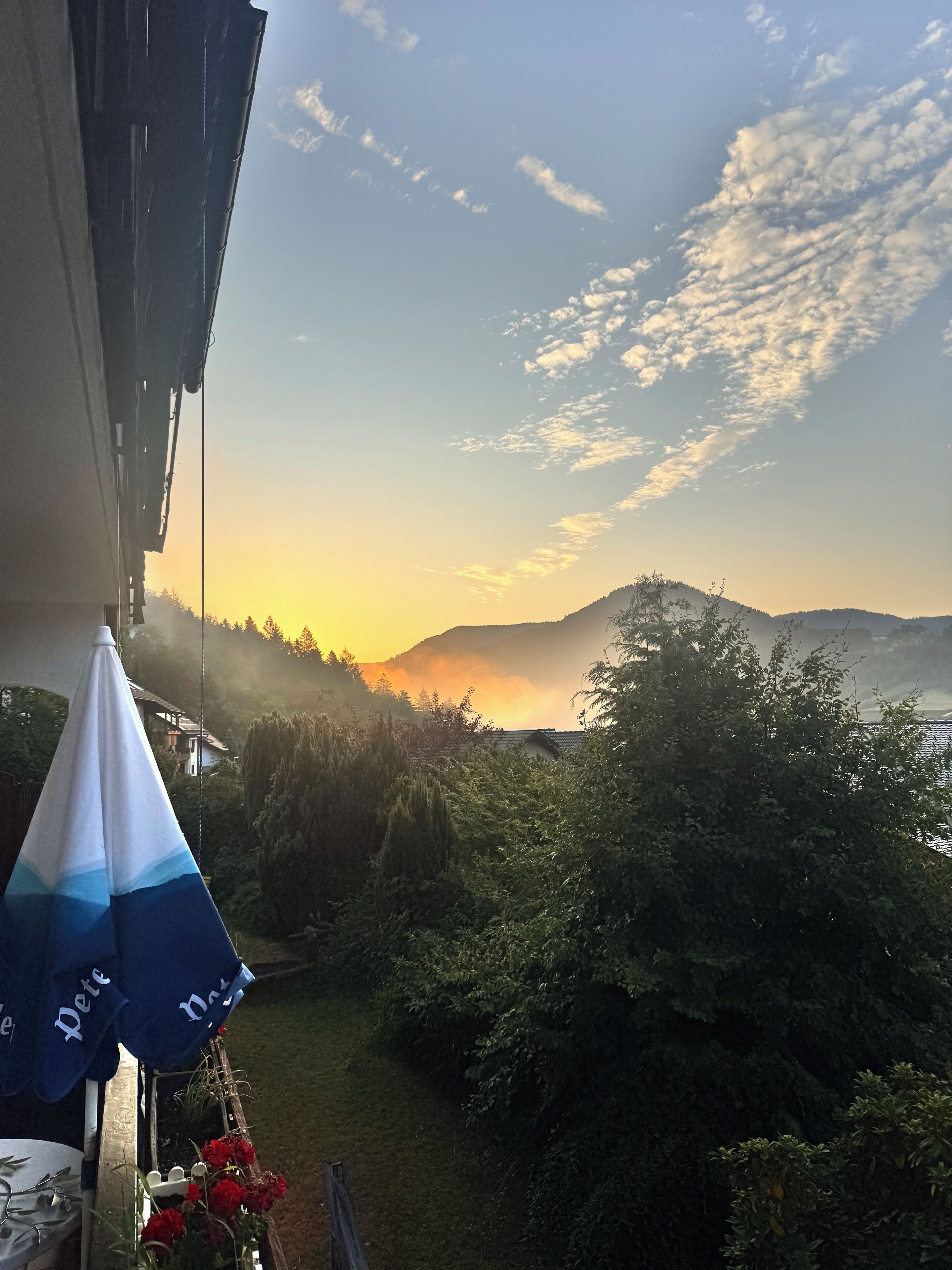 Een uitzicht op een bergachtig landschap met een zonsondergang, een tuin met planten, en een gebouw met een balkon.