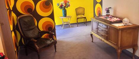 A room with a patterned wall, a wooden chair, a small table with a vase of flowers, and a wooden dresser with drawers.