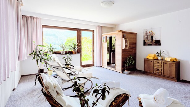 Una habitación amplia con un gran ventanal corredero que da a un balcón, múltiples sillones de descanso, un armario de madera con toallas y varias plantas en macetas.