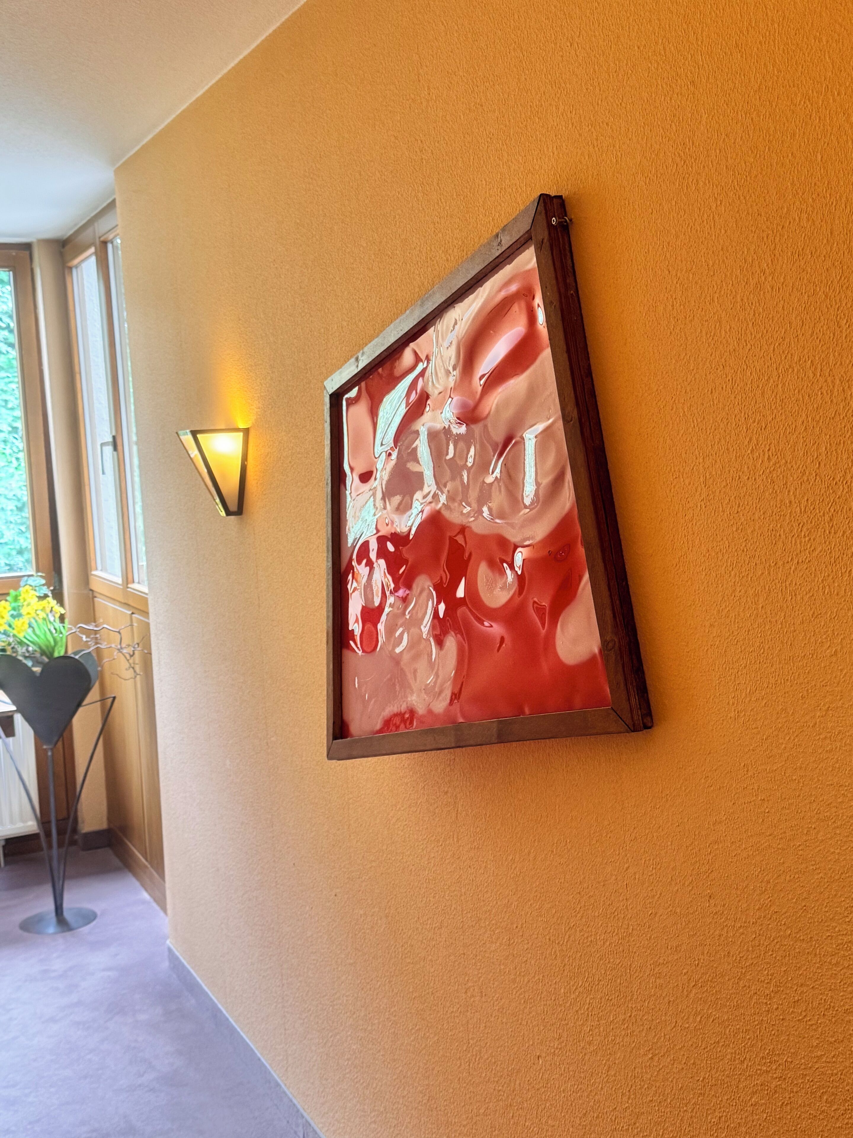 Ein gerahmtes abstraktes Gemälde an einer gelben Wand, eine Wandleuchte und ein Blick in einen Raum mit Fenster und Pflanze.
