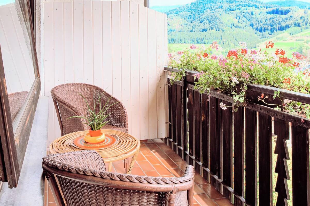 Ein Balkon mit Korbmöbeln, Blick auf eine Berglandschaft und einem orangefarbenen Sonnenschirm.