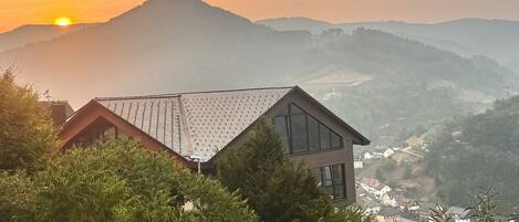 A house with a sloped roof, surrounded by dense greenery and overlooking a valley.
