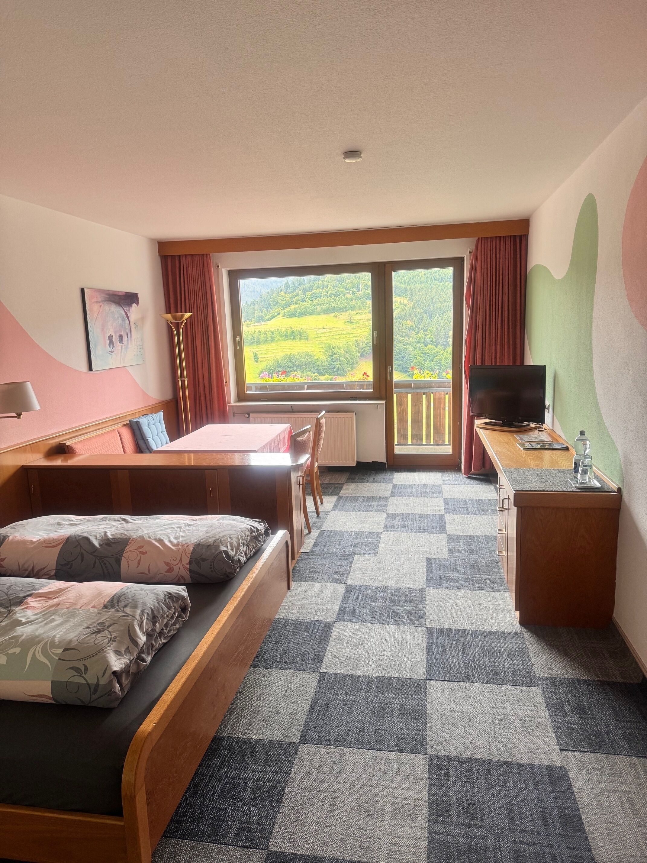 A hotel room with a bed, a desk with a television, a window with a view of green hills, and a painting on the wall.
