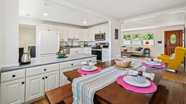 A modern kitchen with white cabinets, a stainless steel refrigerator, and a microwave. There is a dining area with a wooden table, pink placemats, and a colorful woven runner. - Cozy Pet & Family-friendly fully fenced beautiful  backyard (Arvada)