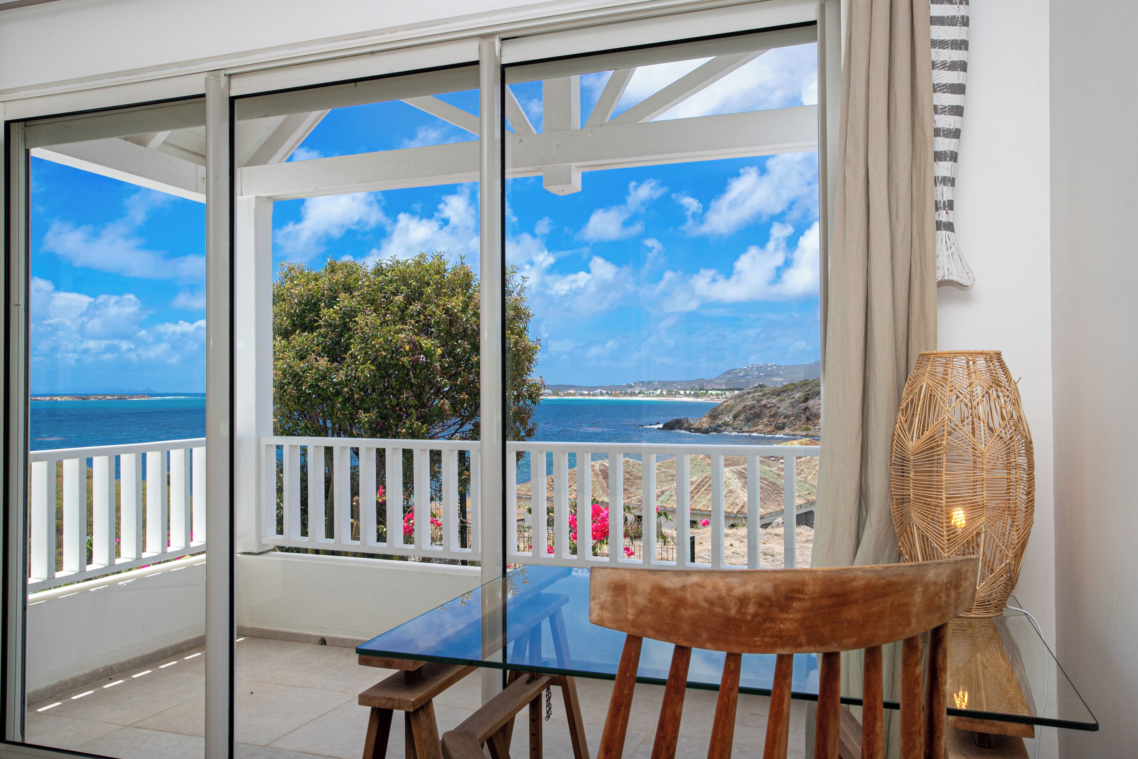A balcony with a glass table and chair, overlooking a coastal view with a clear sky.