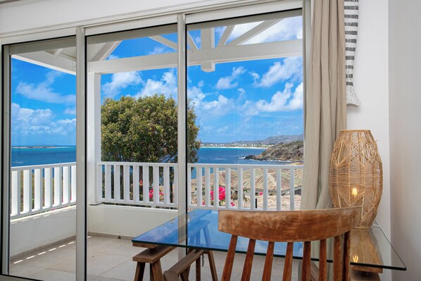 A balcony with a glass table and chair, overlooking a coastal view with a clear sky.