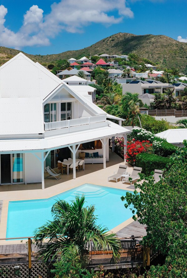 Ein weißes, zweistöckiges Haus mit Pool, umgeben von viel Grün und einem Berg im Hintergrund.