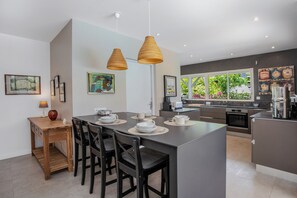 Cocina moderna con una isla central, sillas de comedor, luces colgantes y vistas al exterior a través de amplios ventanales.