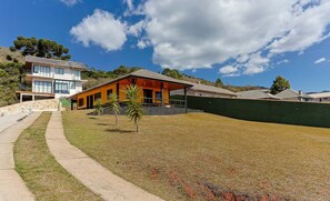 A modern two-story house with a covered patio, a grassy yard, and a hillside backdrop. - Casa 3Q completa c/ Piscina e Churrasq. DOJ0690 (Campos do Jordão)