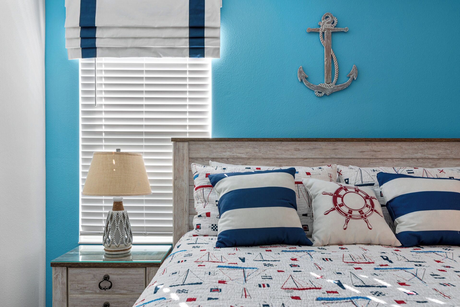 Quarto com tema náutico, apresentando uma cama com roupa de cama listrada e decoração de âncora, um criado-mudo com luminária e uma janela com persianas.
