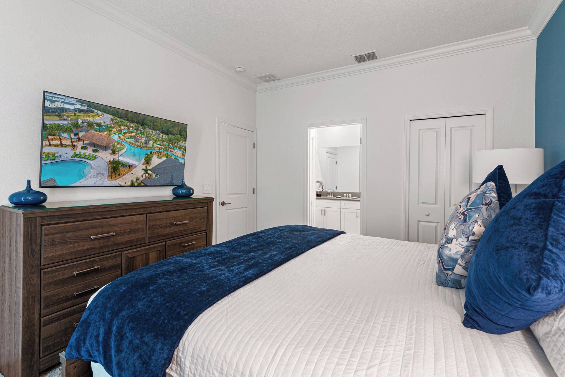 A bedroom with a bed, a dresser, a TV on the wall, and a bathroom visible through an open door.