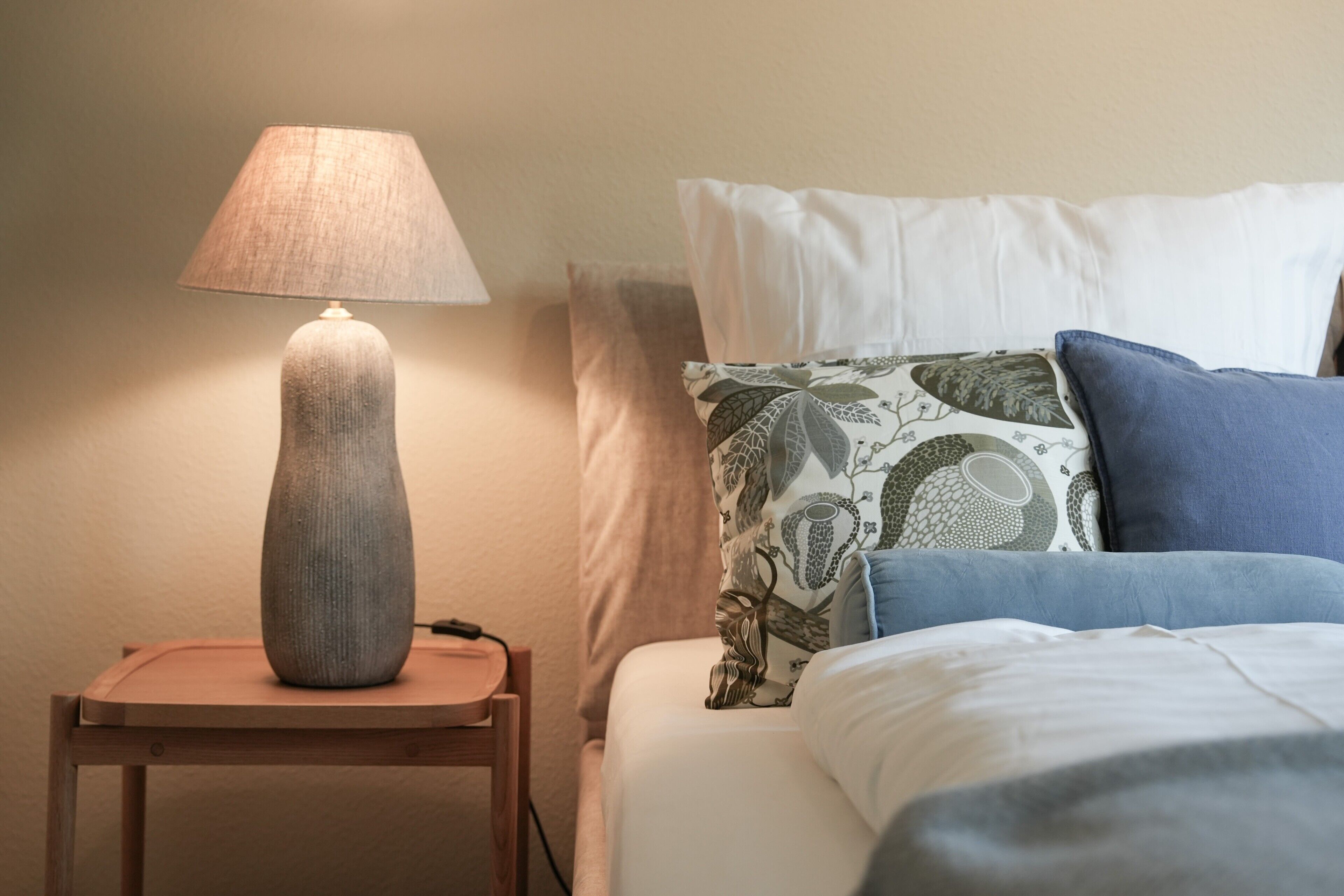 Un lit avec une literie blanche et bleue, un oreiller à motifs et une lampe sur une table de chevet en bois.