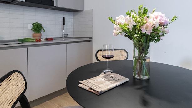 Une cuisine moderne avec des meubles blancs, un plan de travail noir, une plante en pot, un verre à vin et un vase rempli de fleurs, le tout posé sur une table ronde.