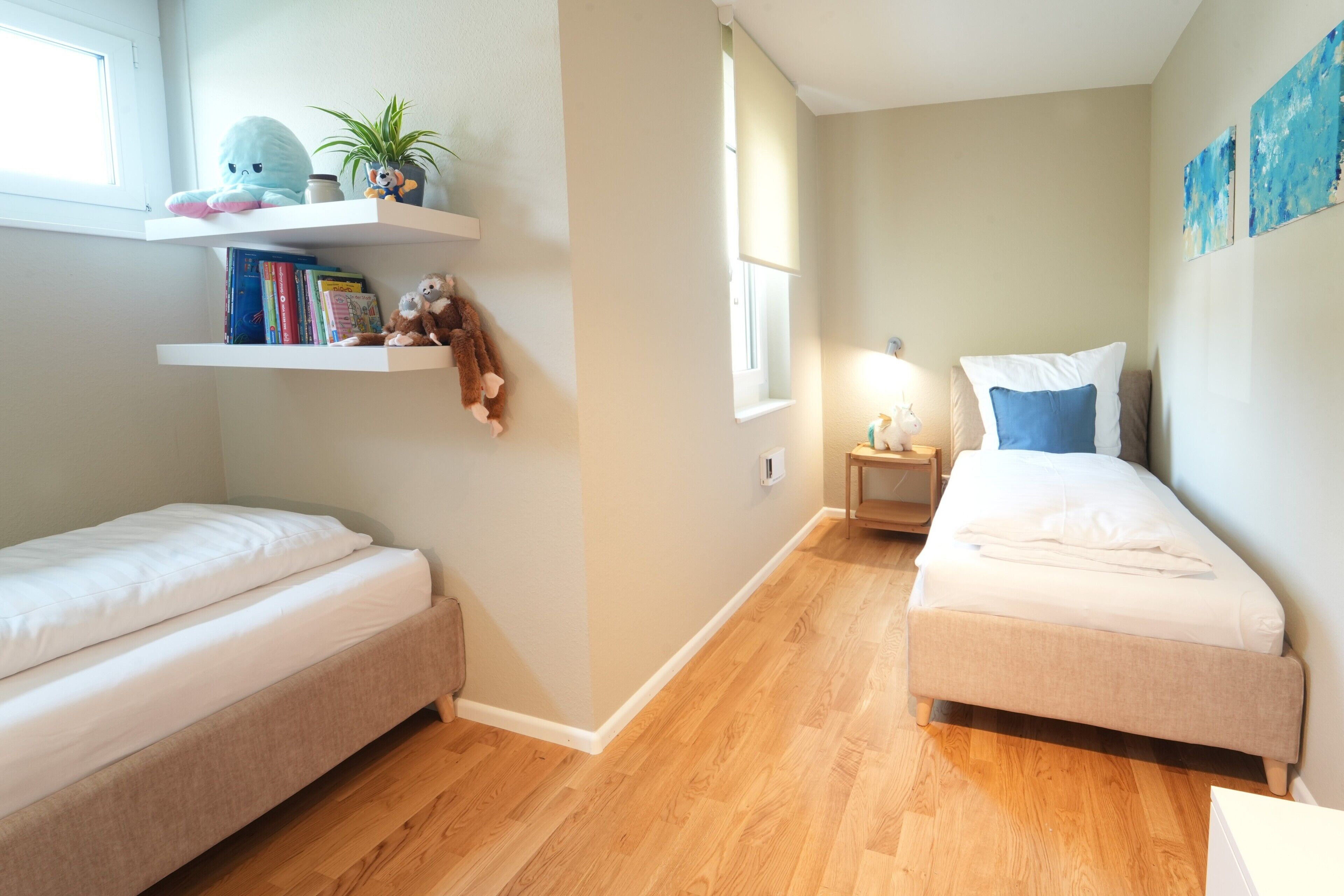 Ein Zimmer mit zwei Betten, Holzboden, einem Fenster, einem Nachttisch mit Lampe sowie Regalen mit Büchern und einem Stofftier.