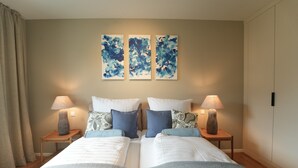 Quarto de hotel com uma cama, duas mesas de cabeceira, luminárias e três obras de arte abstratas nas paredes.