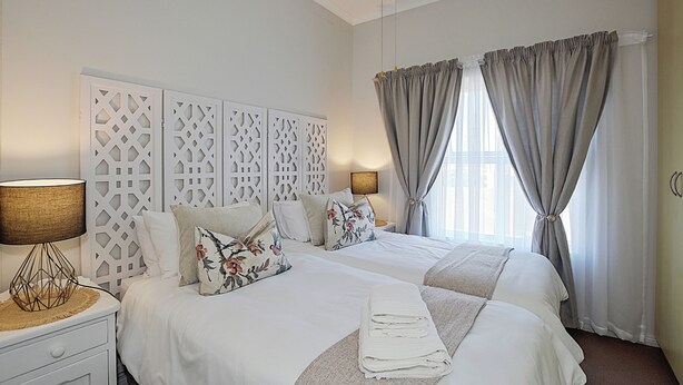 A hotel room with two beds, a decorative headboard, a bedside table with a lamp, and curtains.