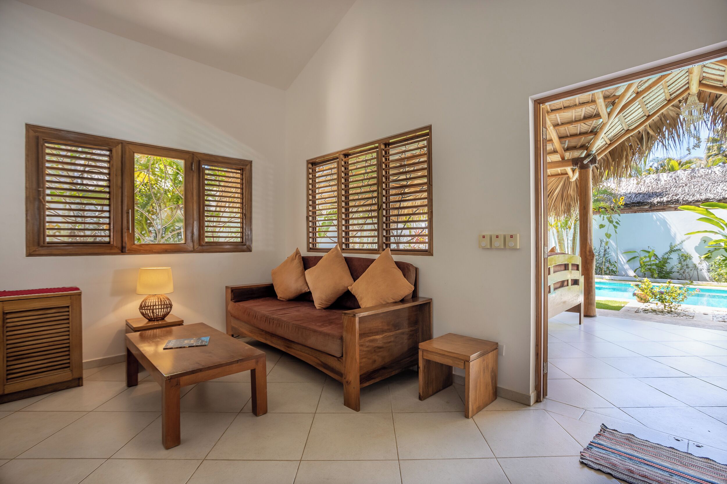 Ein Wohnzimmer mit einer Holzsofa, zwei kleinen Tischen und einer Lampe. Große Fenster mit Holzjalousien, ein Blick auf einen Pool im Freien und ein Dach mit Reet.