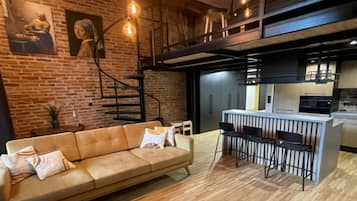 A modern interior with a brick wall, a sofa, a dining area with bar stools, and a staircase.