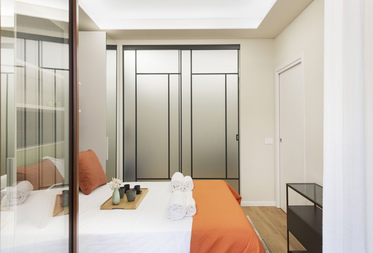 A modern bedroom with a bed, a wooden side table, a mirror, and a wardrobe with sliding doors.