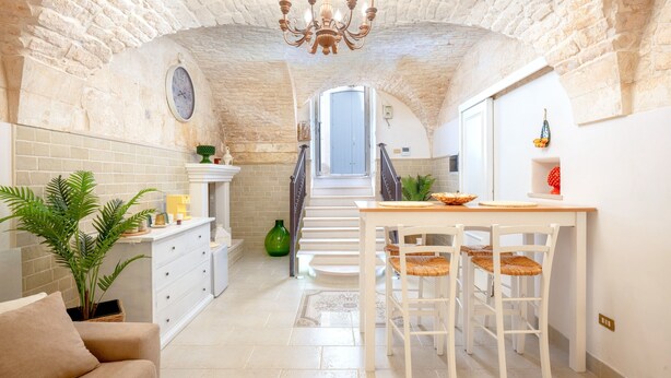 A spacious kitchen with a staircase leading to a balcony, a dining area with white furniture, and a large green plant.