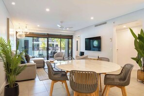 A modern living room with a round table, comfortable seating, a flat-screen TV, and large windows.
