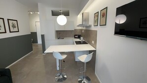 A modern kitchen with a white countertop, two bar stools, a built-in oven, and a flat-screen TV mounted on the wall.