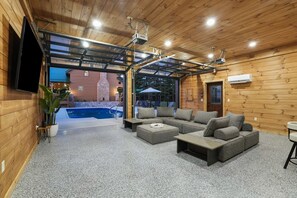 Ein modernes Blockhaus-Interieur mit einer großen Eckcouch, einem flachbildfernseher an der Wand und einem Pool, der durch einen offenen Durchgang sichtbar ist.