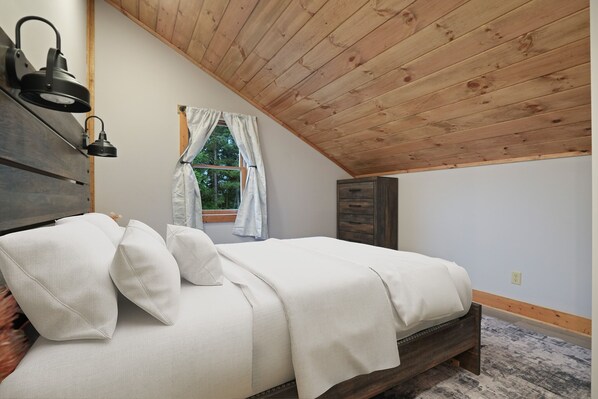 A bedroom with a wooden ceiling, a bed with white bedding, a nightstand with a lamp, and a window with curtains. - The View at Grizzly Mountain | Hocking Hills, Pool (Creola)