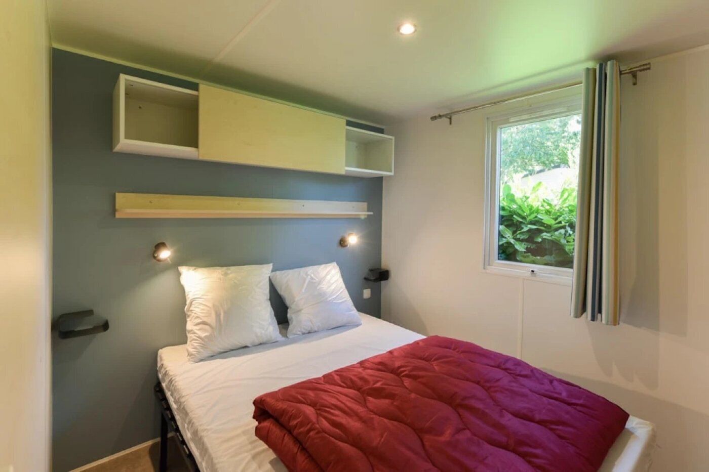 A bedroom with a bed, two pillows, a red blanket, a window with curtains, and built-in shelves above the bed.