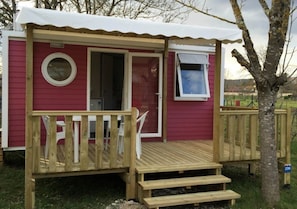 Uma cabana pequena, térrea, com um deck de madeira, uma porta e uma janela.
