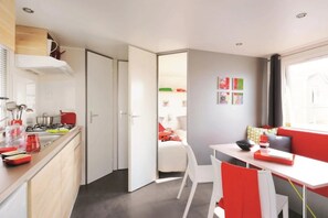 A compact kitchen with a stove, sink, and white cabinets. A dining area with a table and chairs is visible, and a bedroom with a bed and a window is seen through an open door.