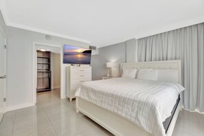 A modern hotel bedroom with a large bed, a TV mounted above the dresser, a walk-in closet, and a view of the sunset through the window. - Suite on the Ocean with City View (Fort Lauderdale)
