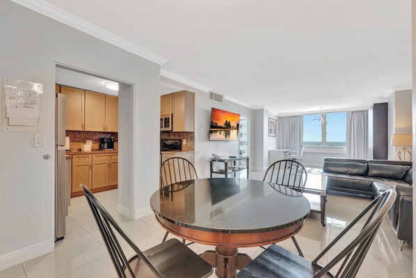 A modern living room with a round glass table, a black leather sofa, and a kitchen in the background. - Suite on the Ocean with City View (Fort Lauderdale)