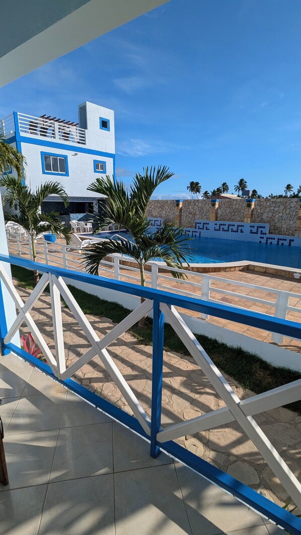 Zona de piscina con muro de piedra, palmeras y un edificio de fachada azul y blanca.