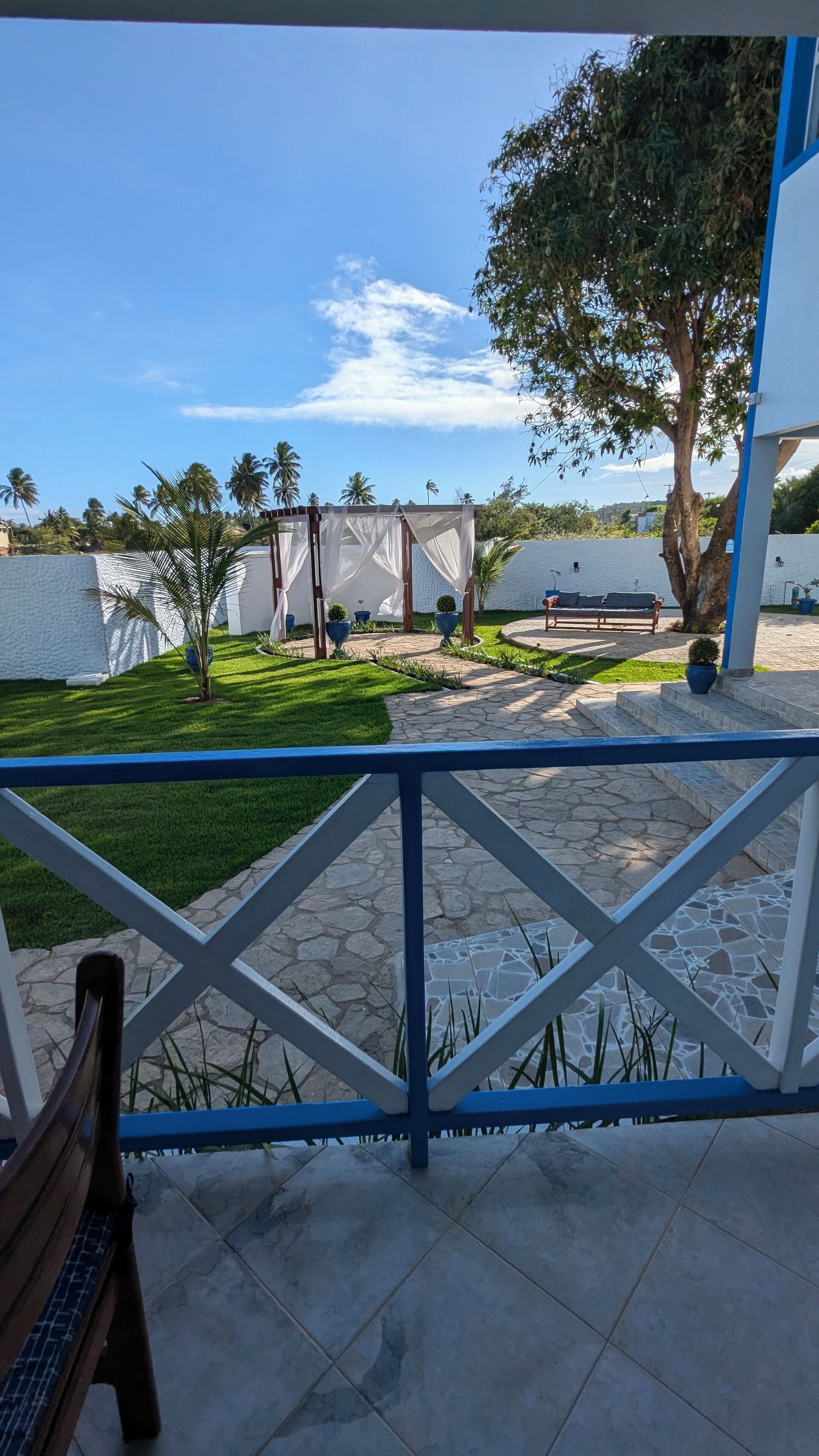 En terrasse med blåt gelænder, en træbænk og en stentrappe, der fører til et siddeområde med en hvid pavillon.