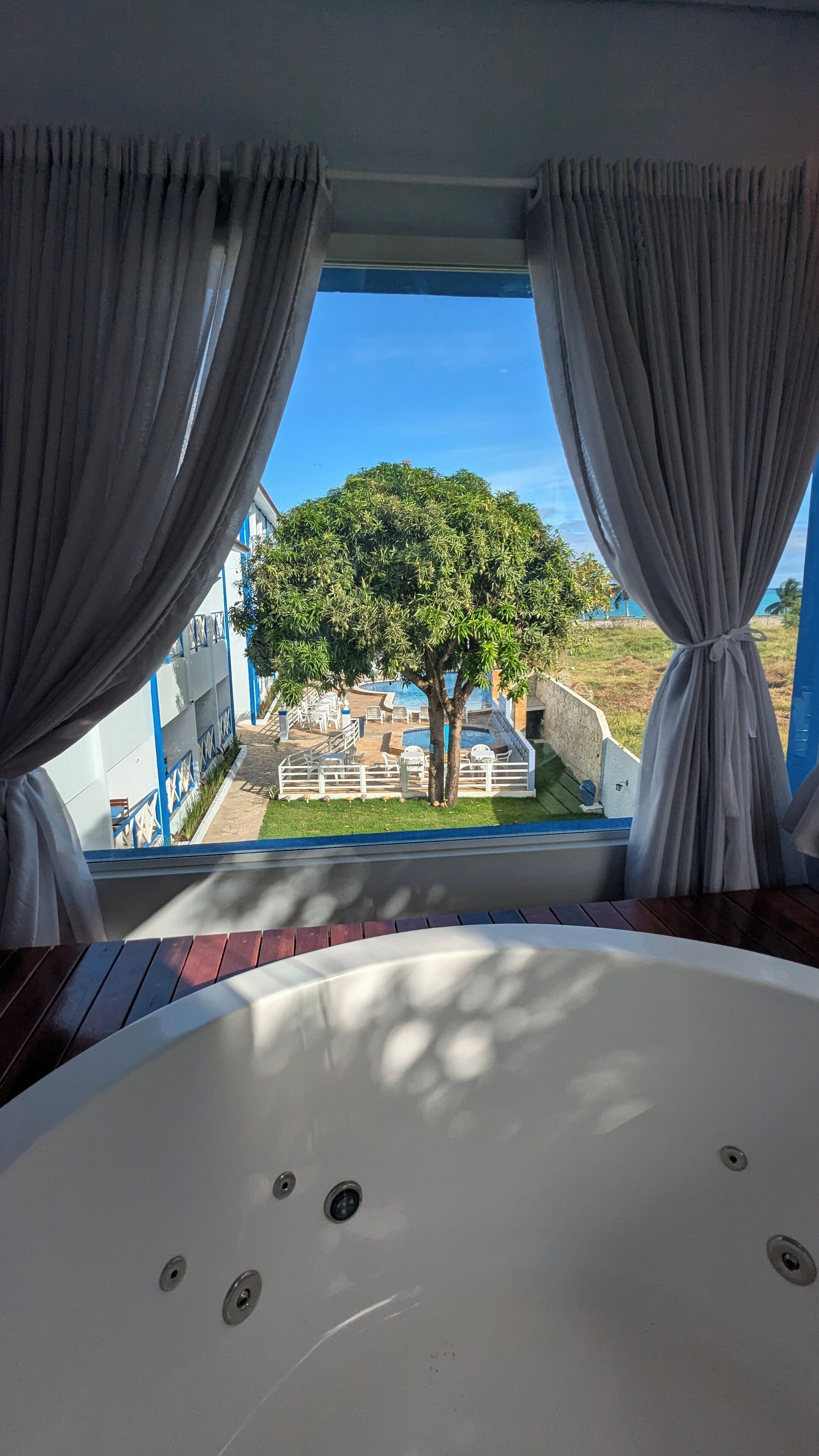 A room with a large bathtub, curtains, and a view of a pool and tree.
