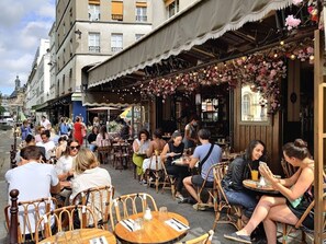 An outdoor café with wooden tables and chairs, surrounded by buildings.