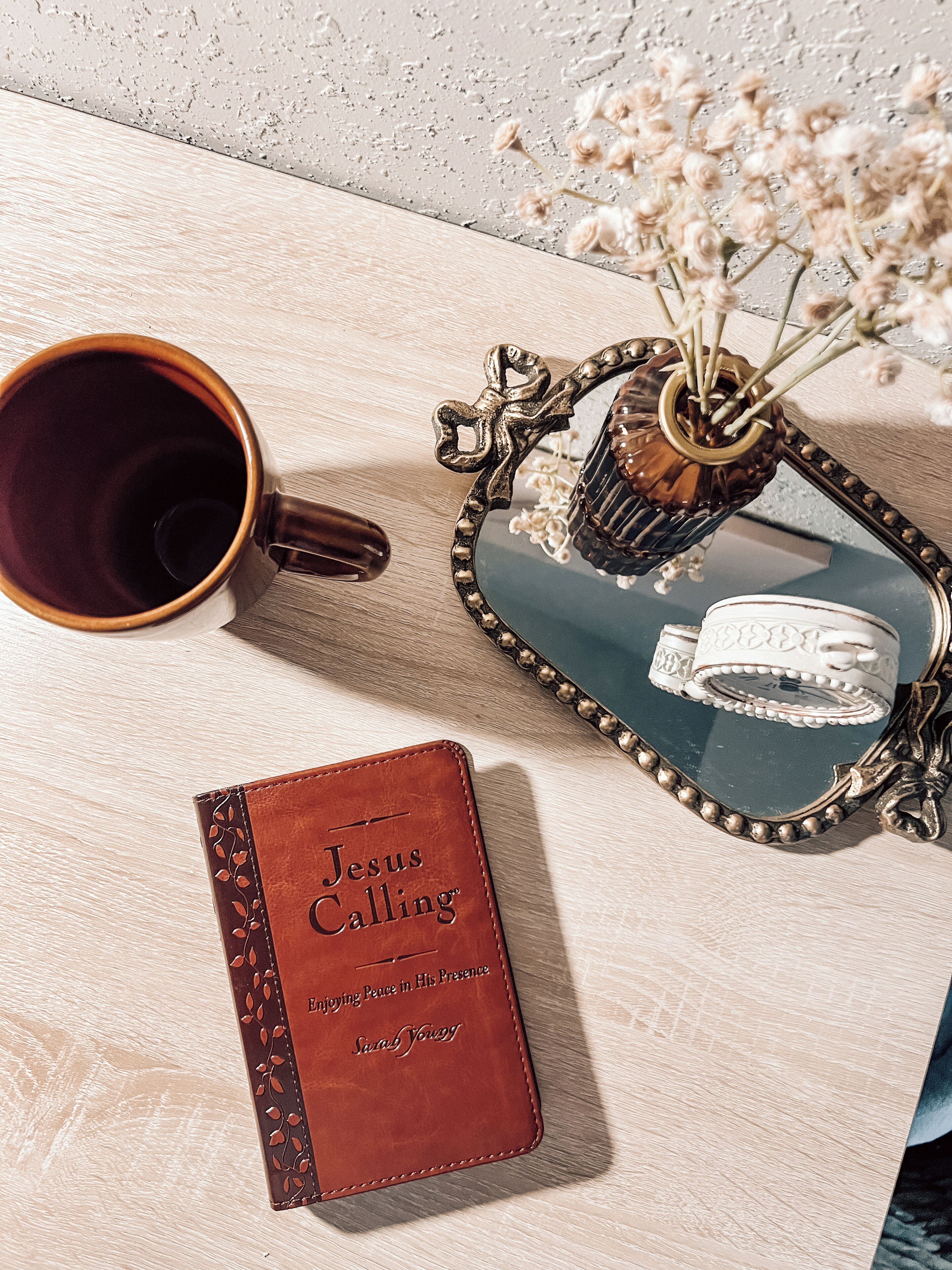 A wooden table with a brown leather journal titled "Jesus Calling," a dark brown mug, and a decorative mirror with a floral vase.